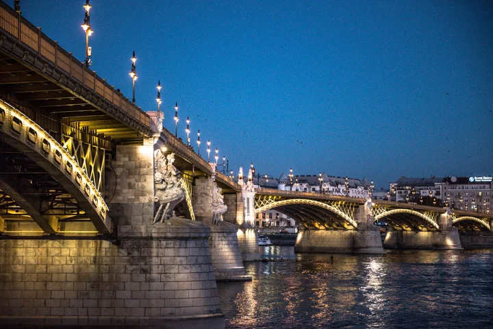 An ornate stone and metal bridge lit above the river at dusk