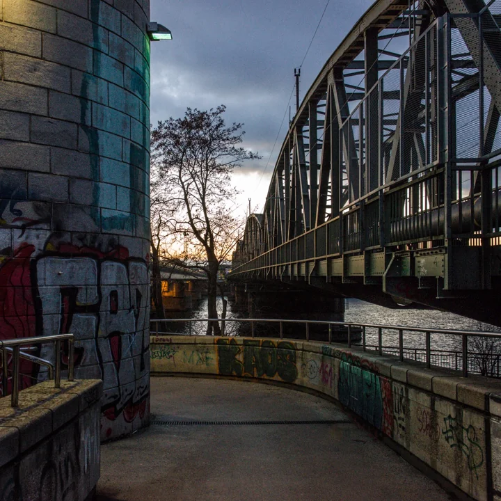 A riverside walkway curving past a graffiti-marked pier and a metal bridge